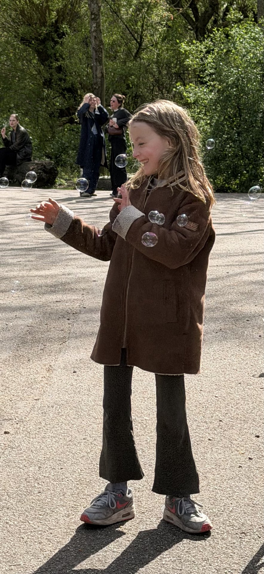 A young girl with long hair, wearing a brown coat and sneakers, joyfully plays with bubbles in Flevopark, Oost, Amsterdam